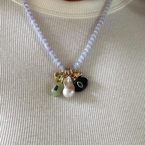 以下が含まれることがあります： 淡いブルーのビーズのネックレス。緑色の石、真珠、黒い石が付いたゴールドのチャームが付いています。白いリブ編みのシャツの上に着用されています。