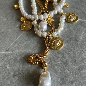 May include: A layered necklace with multiple strands of white pearls and gold chains. The necklace features several gold charms, including eye-shaped pendants with a single diamond accent. The necklace also has a large, teardrop-shaped pearl pendant.