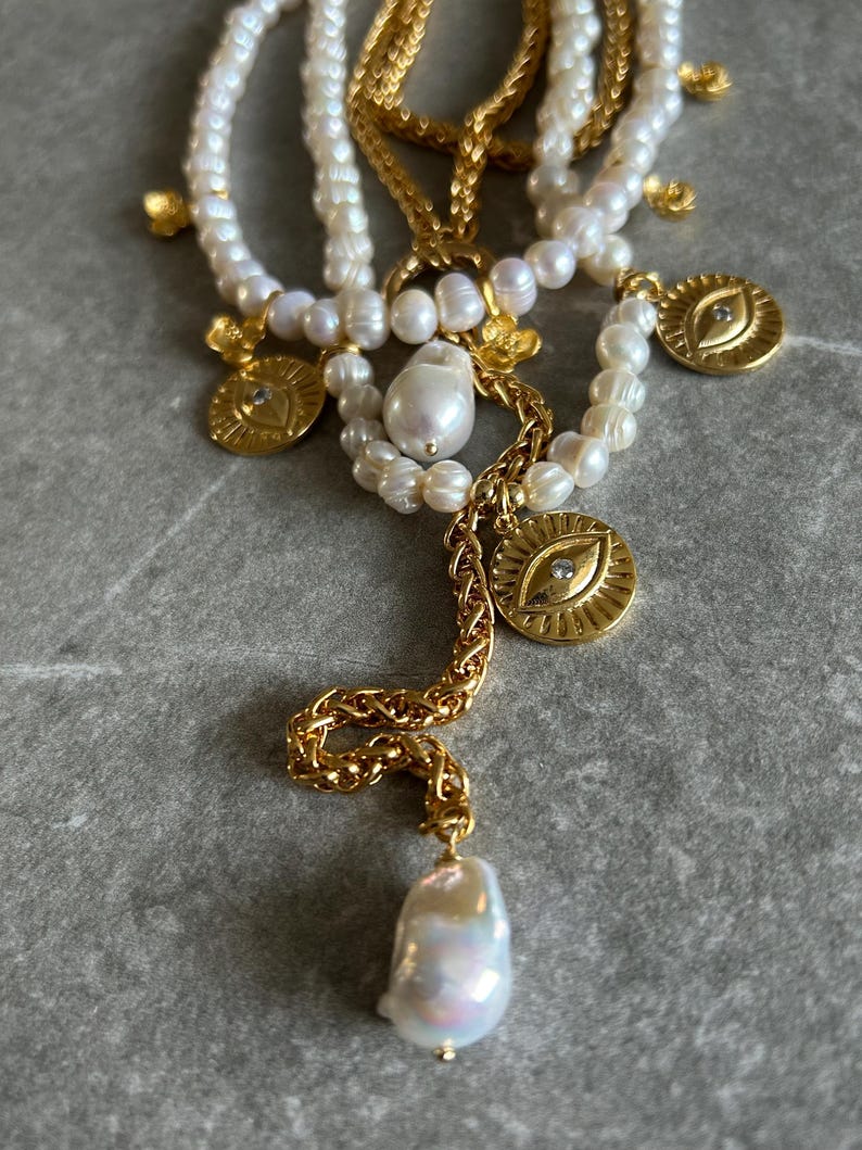 May include: A layered necklace with multiple strands of white pearls and gold chains. The necklace features several gold charms, including eye-shaped pendants with a single diamond accent. The necklace also has a large, teardrop-shaped pearl pendant.
