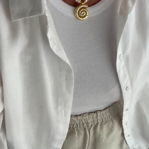 May include: A white tank top with a gold necklace featuring a round pendant with a clear stone. The tank top is layered under a white button-down shirt. The person is wearing beige linen shorts.