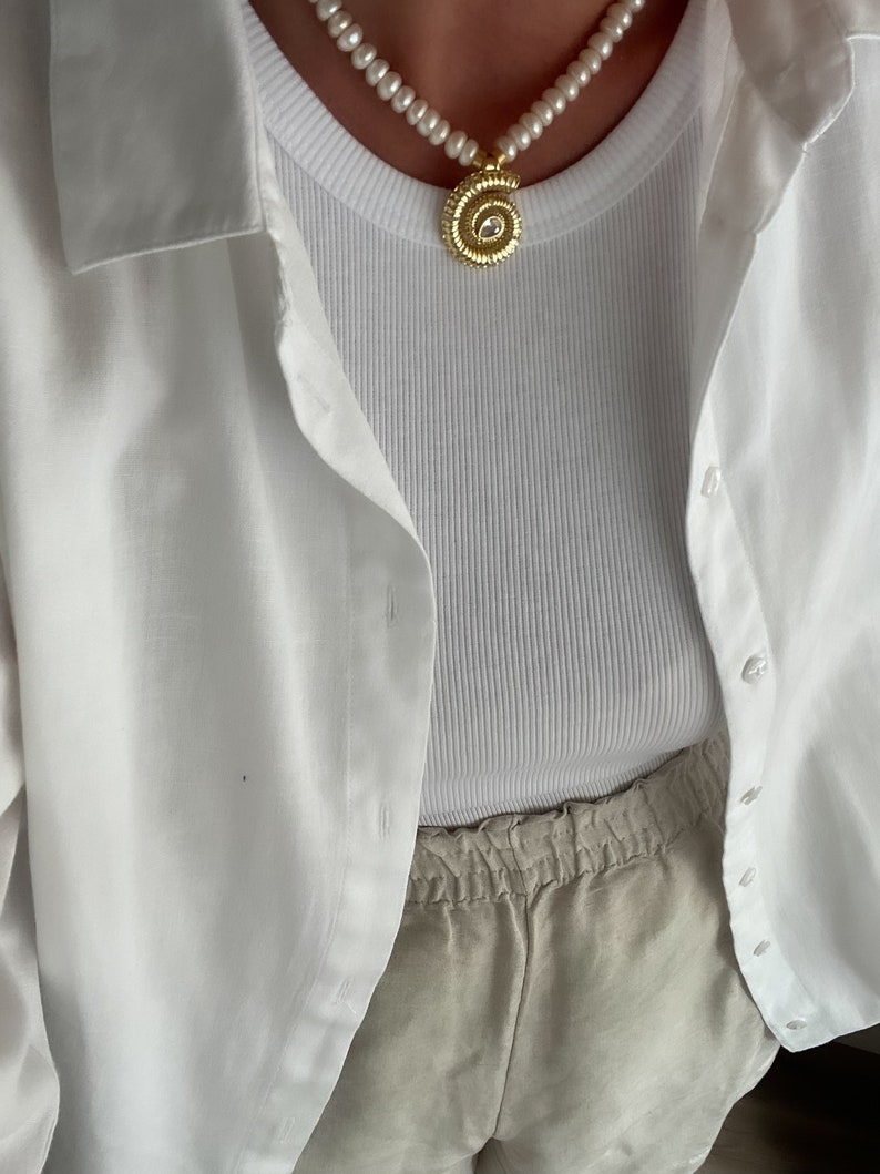May include: A white tank top with a gold necklace featuring a round pendant with a clear stone. The tank top is layered under a white button-down shirt. The person is wearing beige linen shorts.