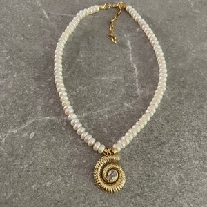 May include: A white pearl necklace with a gold spiral pendant featuring a clear rhinestone. The necklace is on a gray background.