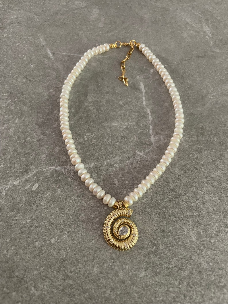May include: A white pearl necklace with a gold spiral pendant featuring a clear rhinestone. The necklace is on a gray background.