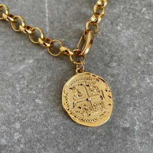 May include: A gold chain necklace with a large, round, gold coin pendant featuring a detailed design of a cross with four symbols.