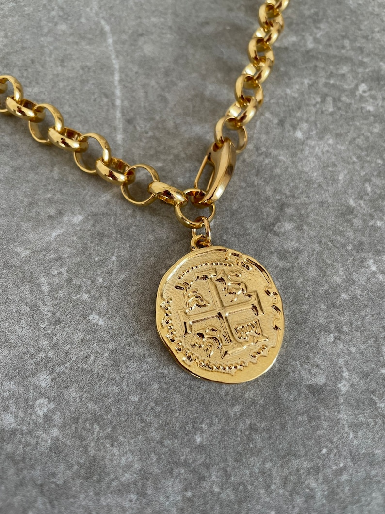 May include: A gold chain necklace with a large, round, gold coin pendant featuring a detailed design of a cross with four symbols.