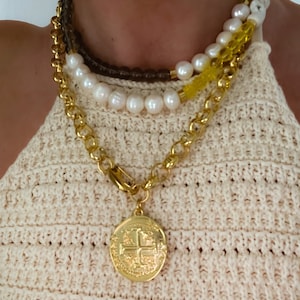 May include: Layered gold chain necklace with a large gold coin pendant featuring a cross design. The necklace is worn over a white pearl and brown beaded necklace.