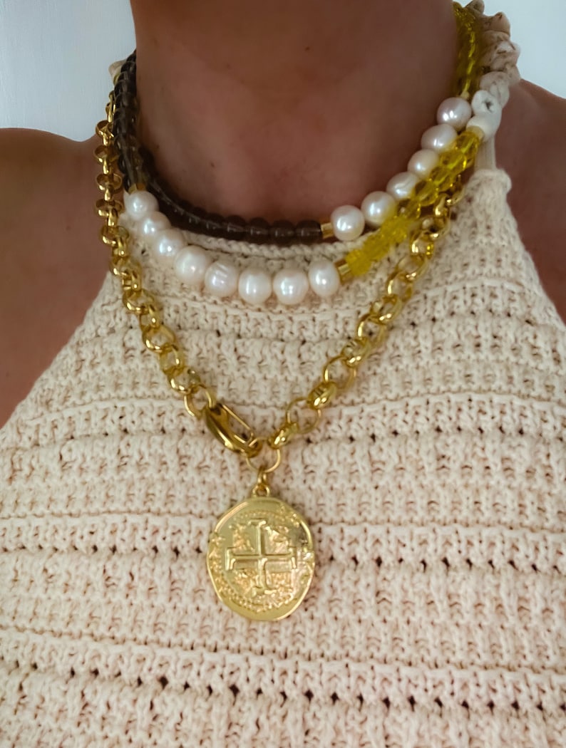 May include: Layered gold chain necklace with a large gold coin pendant featuring a cross design. The necklace is worn over a white pearl and brown beaded necklace.