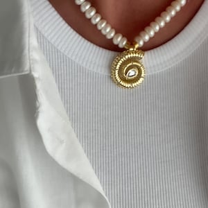 May include: A white pearl necklace with a gold spiral pendant featuring a clear gemstone. The necklace is worn by a person wearing a white shirt.