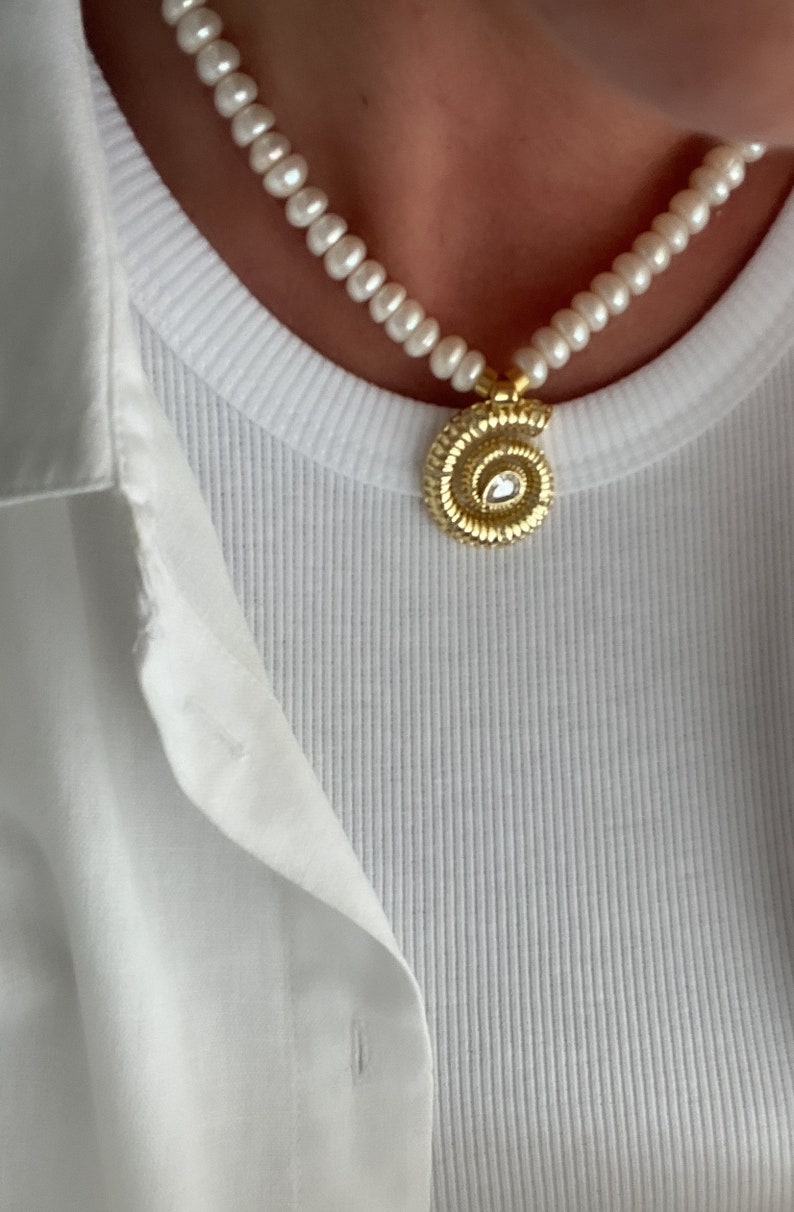 May include: A white pearl necklace with a gold spiral pendant featuring a clear gemstone. The necklace is worn by a person wearing a white shirt.