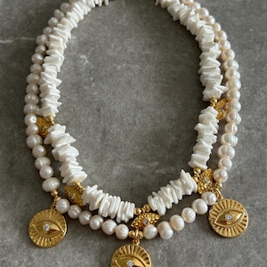May include: A layered necklace featuring white shell chips and pearls. Gold-toned pendants with an eye design and a small diamond are suspended from the necklace. The necklace has a gold-toned clasp.