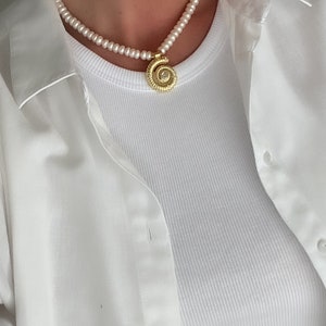 May include: A white pearl necklace with a gold pendant featuring a spiral design. The necklace is worn by a person wearing a white shirt.
