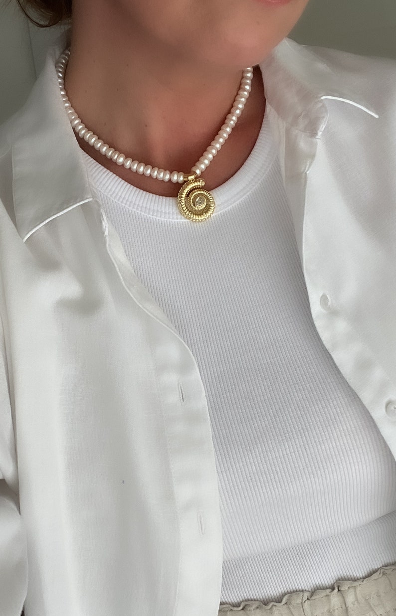 May include: A white pearl necklace with a gold pendant featuring a spiral design. The necklace is worn by a person wearing a white shirt.