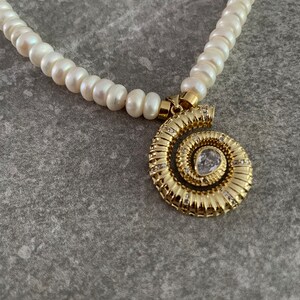 May include: A white pearl necklace with a gold spiral pendant featuring a clear rhinestone. The pendant is detailed with small, sparkling rhinestones.