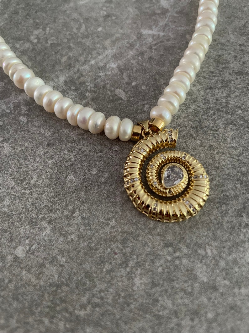 May include: A white pearl necklace with a gold spiral pendant featuring a clear rhinestone. The pendant is detailed with small, sparkling rhinestones.