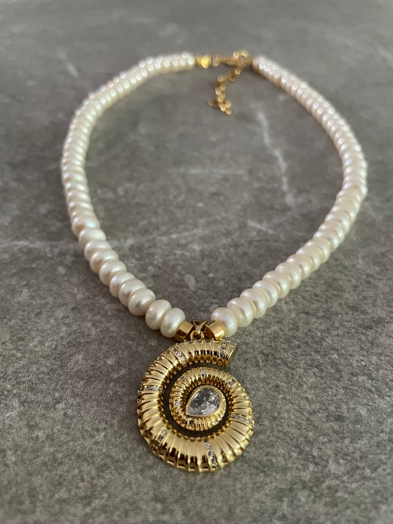 May include: A white pearl necklace with a gold spiral pendant featuring a clear rhinestone. The pendant is detailed with small gold accents.
