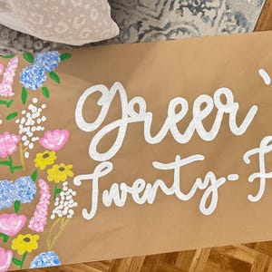 May include: A brown paper banner with the text "Greer's Twenty-First" in white script. The banner is decorated with painted flowers in pink, blue, yellow, and white, arranged in a floral border.