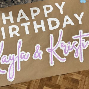 May include: A brown paper banner with the words "HAPPY BIRTHDAY" in white block letters. Below, the names "Kayla & Kristi" are written in a light purple script with white outlines. Decorative flowers in light blue and purple are scattered around the text.