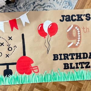 May include: A brown paper banner with a football theme. It features a football, goal posts, a red helmet, and green grass at the bottom. The banner reads "Jack's 9th Birthday Blitz" with red and white balloons and pennants.