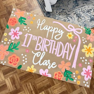 May include: A hand-painted birthday sign on brown paper, featuring the text "Happy 17th Birthday Clare" in white and pink. The sign is decorated with colorful flowers and green leaves.