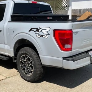May include: A white pickup truck with a black toolbox in the bed. The truck features a black and white "FXR 4x4" decal on the side. The truck has a black tyre with the word "Goodyear" on the tyre.