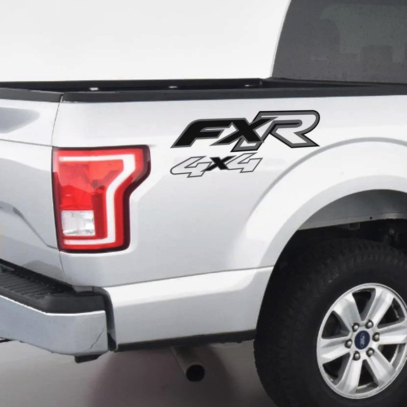 May include: A white truck bed with a black and grey "FXR 4x4" decal. The decal is on the side of the truck bed, near the rear tyre and taillight. The taillight is red and white.