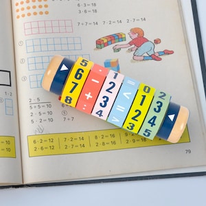 Könnte beinhalten: Ein farbenfrohes Holzspielzeug mit drehbaren Zahlenrädern und mathematischen Symbolen. Das Spielzeug soll Kindern helfen, grundlegende mathematische Konzepte zu lernen. Das Spielzeug ist auf einem aufgeschlagenen Buch mit mathematischen Aufgaben abgebildet.