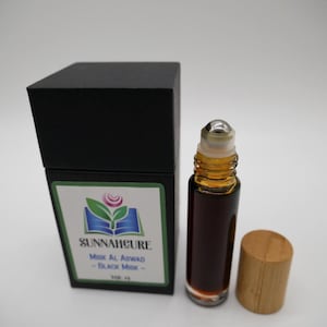 May include: A roll-on bottle of dark liquid with a silver rollerball top and a wooden cap, next to a black box with a green and white label. The label reads "SUNNAHCORE Musk Al Aswad - Black Musk -".