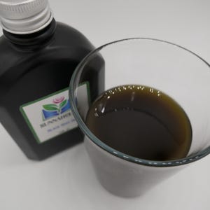 May include: A black bottle with a silver cap and a clear glass filled with a dark brown liquid. The bottle has a green and white label with a floral design and text. The glass is next to the bottle.