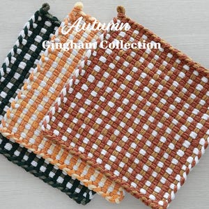 May include: Three square woven potholders featuring a gingham pattern in dark green, orange, and rust brown, each combined with white. The text Autumn Gingham Collection is printed in white over the center of the three stacked textile items.