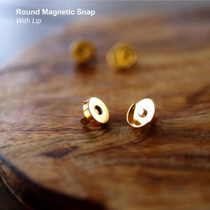 May include: Close-up of gold-colored round magnetic snaps with a lip, displayed on a wooden surface. The text "Round Magnetic Snap With Lip" is visible in the image. These snaps are likely used for bags or other craft projects.