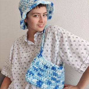 Puede incluir: Un sombrero de cubo de crochet blanco y azul con un ala ancha. El sombrero lo lleva una persona que lleva una camisa blanca abotonada con un estampado de cachemira. La persona tambi&eacute;n lleva un bolso de crochet blanco y azul.