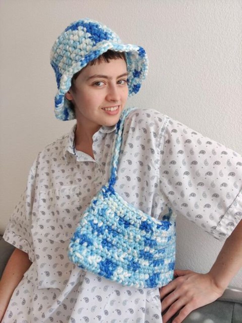 Puede incluir: Un sombrero de cubo de crochet blanco y azul con un ala ancha. El sombrero lo lleva una persona que lleva una camisa blanca abotonada con un estampado de cachemira. La persona tambi&eacute;n lleva un bolso de crochet blanco y azul.