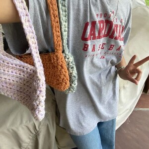 Puede incluir: Tres bolsas de crochet en diferentes colores: lavanda, naranja y verde. Las bolsas cuelgan del brazo de una persona que lleva una camiseta gris con el texto "PROUD CARDINAL BASEBALL" impreso en ella.