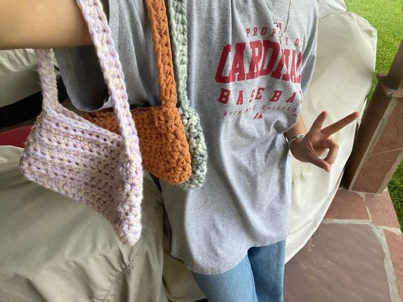 Puede incluir: Tres bolsas de crochet en diferentes colores: lavanda, naranja y verde. Las bolsas cuelgan del brazo de una persona que lleva una camiseta gris con el texto "PROUD CARDINAL BASEBALL" impreso en ella.