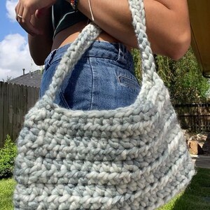 Puede incluir: Un bolso de hombro de crochet gris y blanco con un dise&ntilde;o holgado. El bolso se lleva sobre el hombro de una persona que lleva un top verde sin mangas y pantalones vaqueros azules.