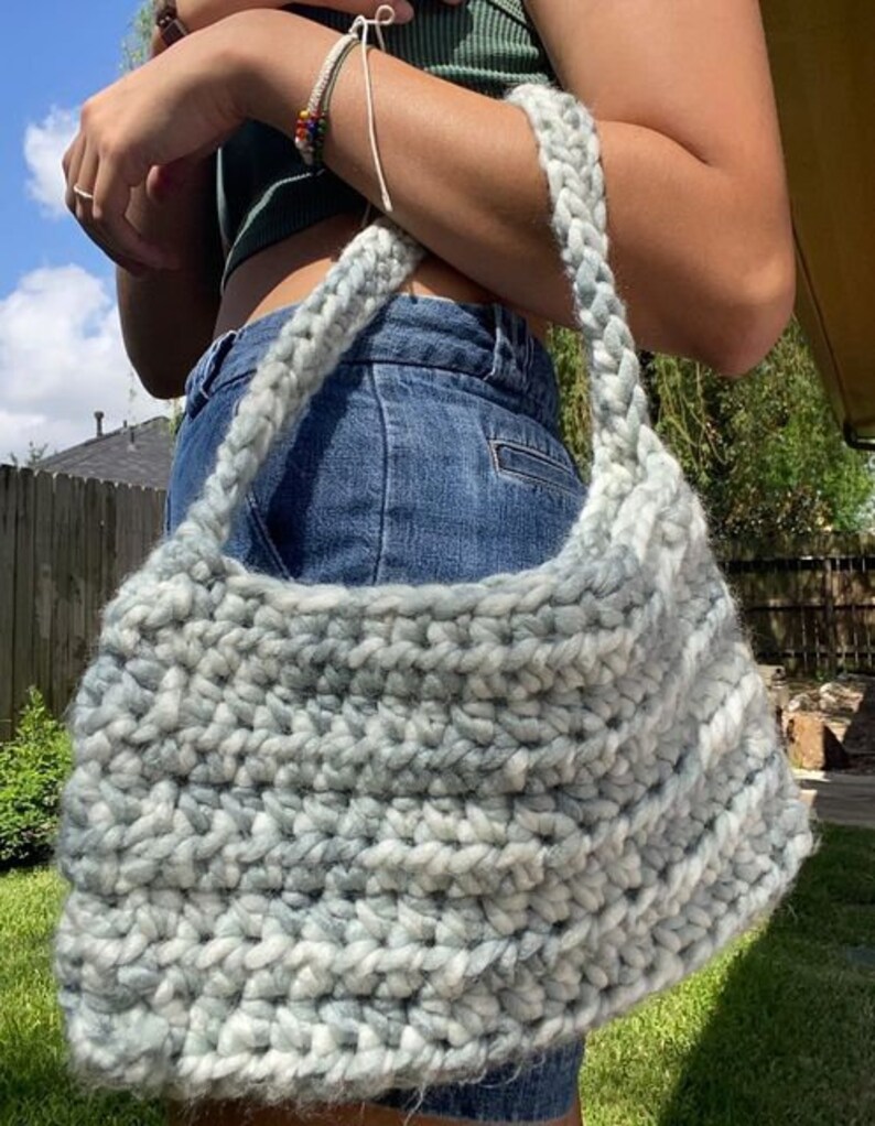 Puede incluir: Un bolso de hombro de crochet gris y blanco con un dise&ntilde;o holgado. El bolso se lleva sobre el hombro de una persona que lleva un top verde sin mangas y pantalones vaqueros azules.