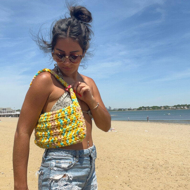 Puede incluir: Una mujer con un top de bikini y pantalones cortos de mezclilla est&aacute; sosteniendo una bolsa de playa de crochet con rayas amarillas, azules y rosas. La bolsa tiene una correa larga y se lleva sobre el hombro.