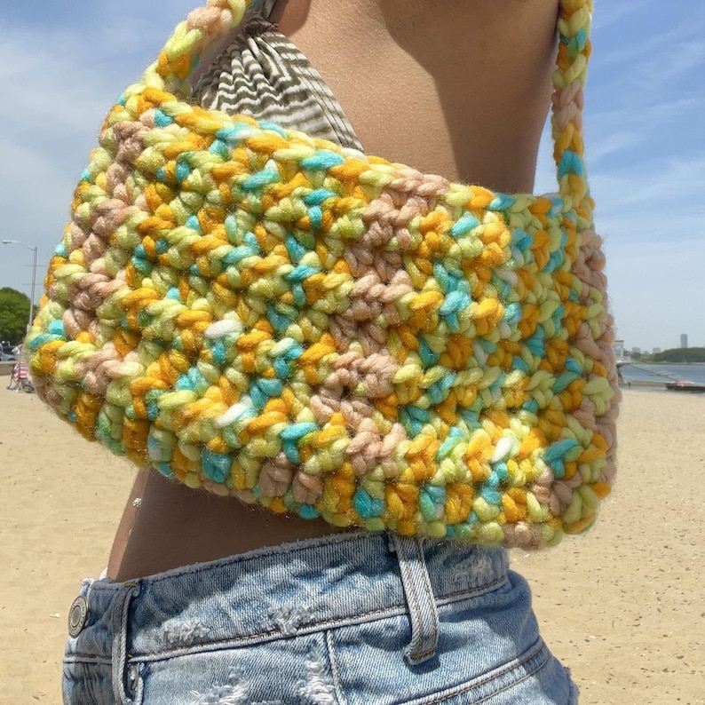 Puede incluir: Un bolso de hombro de crochet con un patr&oacute;n colorido de hilo amarillo, azul y rosa. El bolso se lleva sobre el hombro de una persona que lleva pantalones vaqueros azules.