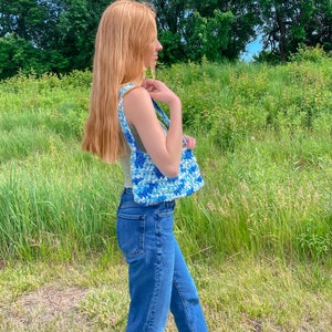 Puede incluir: Una mujer con jeans azules y un top de tirantes marr&oacute;n claro lleva un bolso de crochet azul y blanco.