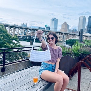 Puede incluir: Un bolso de mano blanco de crochet con una correa larga, sostenido por una persona que lleva un top de crochet morado, pantalones cortos de mezclilla y gafas de sol. La persona est&aacute; sentada en una mesa de madera en una azotea con una vista de la ciudad en el fondo.