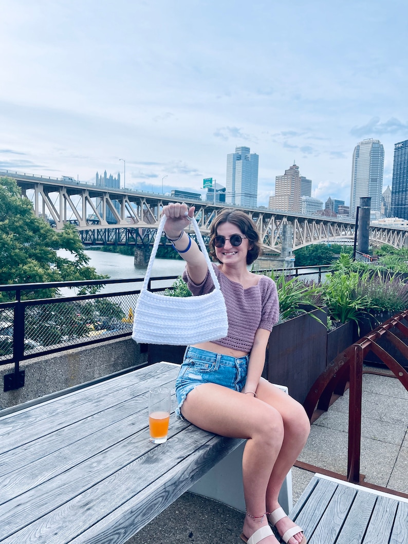Puede incluir: Un bolso de mano blanco de crochet con una correa larga, sostenido por una persona que lleva un top de crochet morado, pantalones cortos de mezclilla y gafas de sol. La persona est&aacute; sentada en una mesa de madera en una azotea con una vista de la ciudad en el fondo.