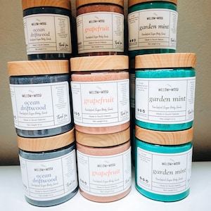 May include: Nine jars of exfoliating sugar body scrub in three scents: ocean driftwood, grapefruit, and garden mint. Each jar has a wooden lid and a label with the scent and brand name "Willow+Wood".