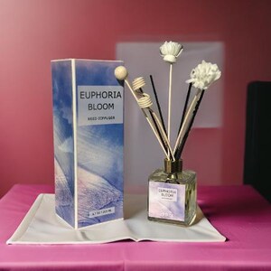 May include: A white and blue reed diffuser bottle with the text "Euphoria Bloom Reed Diffuser" on the label. The bottle is surrounded by white decorative sticks and flowers.