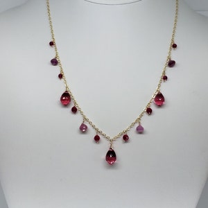 May include: A delicate gold necklace featuring a series of small, round, and teardrop-shaped pink and red gemstones. The necklace is displayed on a white mannequin, showcasing its elegant design and color palette.