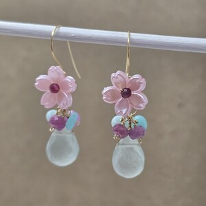 May include: A pair of dangle earrings featuring pink flower-shaped charms with a small red gem in the center. Below the flowers are clusters of small purple and blue beads, and a teardrop-shaped, pale green stone.