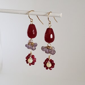 May include: A pair of dangle earrings with red and purple gemstone beads. The earrings feature a teardrop-shaped red gemstone, followed by a cluster of purple beads, and a red flower-shaped bead with a white center.