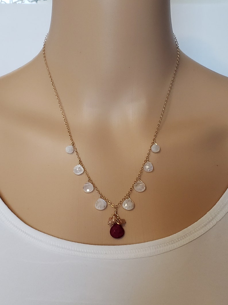 Op de afbeelding: Een gouden ketting met een rode edelsteen hanger en witte traanvormige stenen.