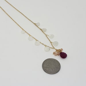 Op de afbeelding: Een gouden ketting met een cluster van kleine witte stenen en een grotere rode steen aan het einde. De ketting is ongeveer zo groot als een munt van 25 cent.