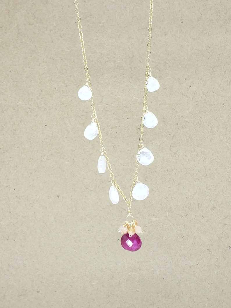 Op de afbeelding: Een gouden ketting met een cluster van kleine witte stenen en een grote roze edelsteen hanger.