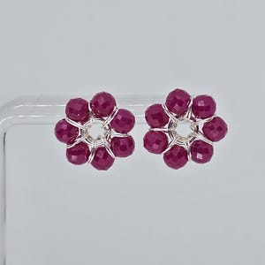 May include: A pair of flower-shaped stud earrings. Each earring features a cluster of faceted, deep magenta beads arranged around a central silver-toned wire. The earrings are set against a plain white background.